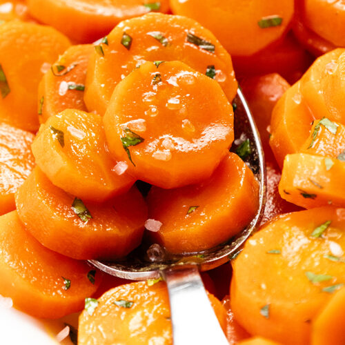 A close-up of easy glazed carrots showcases slices adorned with chopped herbs and coarse salt. A silver spoon cradles some of the glistening orange slices, capturing the vibrant colors and textures of this simple stovetop glazed carrots recipe.
