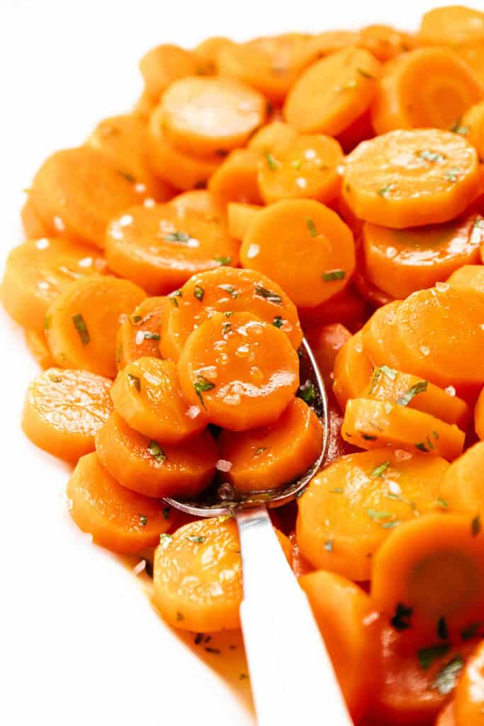 Close-up of glazed carrot slices, an easy stove top dish, garnished with herbs and coarse salt on a white plate with a metal spoon. The bright, glossy carrots beautifully contrast with the fresh green herbs.
