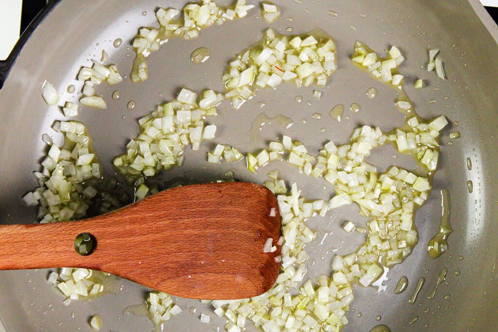 Chopped onions sautéing in oil in a metal pan, being stirred with a wooden spatula—the perfect start for smoky tacos or a flavorful homemade seasoning.