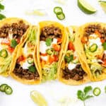 Four ground beef tacos with homemade seasoning, topped with lettuce, diced tomatoes, shredded cheese, sour cream, jalapeños, and cilantro on yellow corn tortillas. Served with lime wedges and red sauce on a white surface.