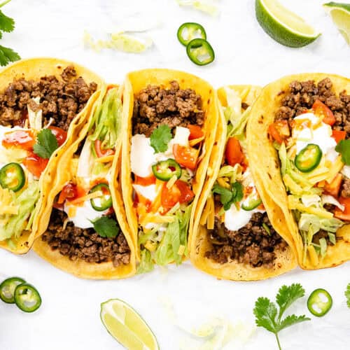Four ground beef tacos with homemade seasoning, topped with lettuce, diced tomatoes, shredded cheese, sour cream, jalapeños, and cilantro on yellow corn tortillas. Served with lime wedges and red sauce on a white surface.