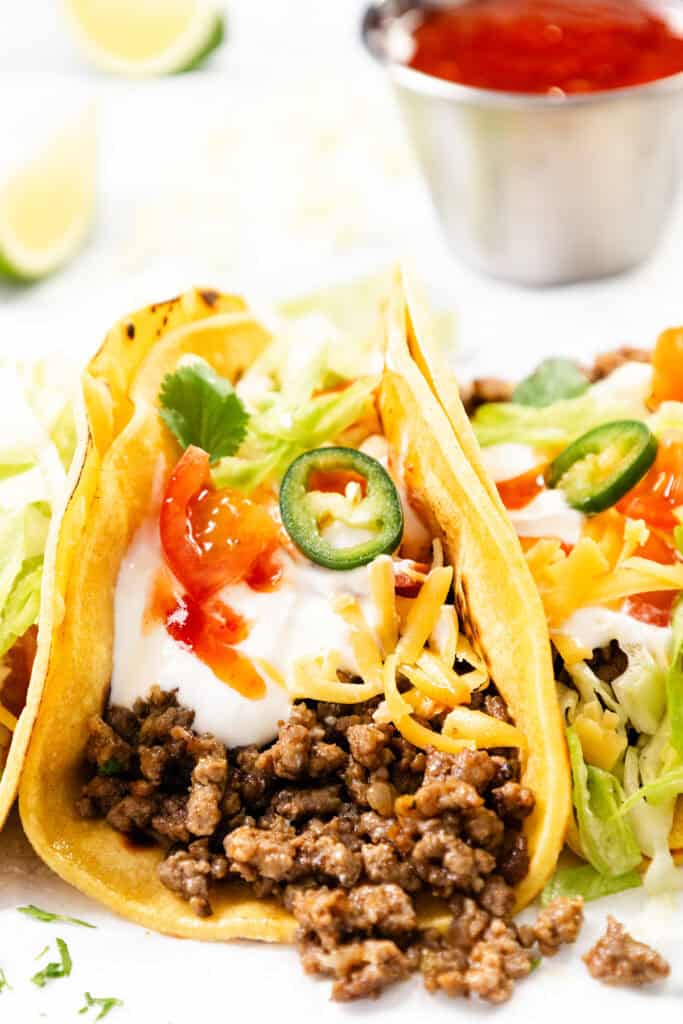 Two hard shell ground beef tacos filled with homemade seasoning, shredded cheese, lettuce, sour cream, jalapeño slices, cilantro, and salsa. A small metal cup of red sauce and lime wedges are in the background.