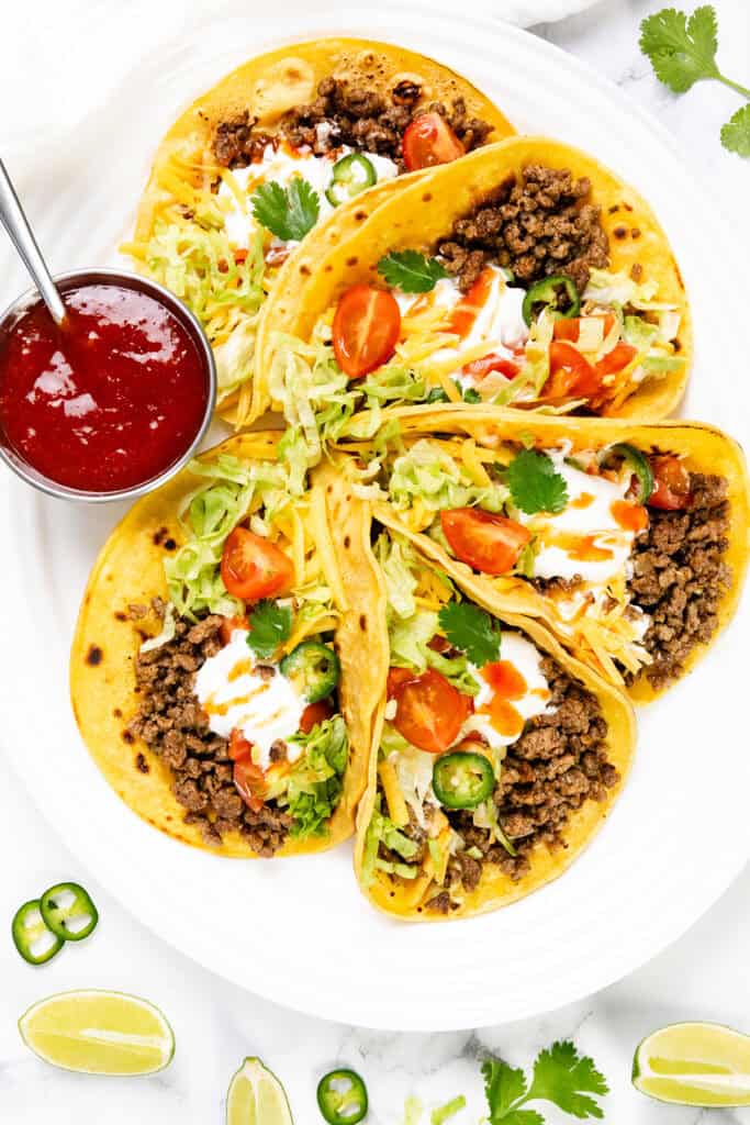 A white plate with four smoky ground beef tacos topped with shredded lettuce, cheese, sliced jalapeños, cherry tomatoes, cilantro, and sour cream. Served with a small cup of red salsa and lime wedges—perfect with homemade seasoning.