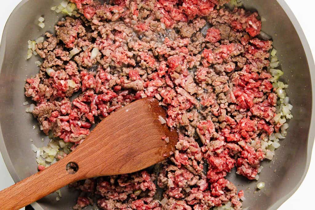 Ground beef and chopped onions sauté in a skillet with a wooden spatula, as the meat turns from raw to browned—perfect as the savory base for Ground Beef Tacos or adding your favorite homemade seasoning.