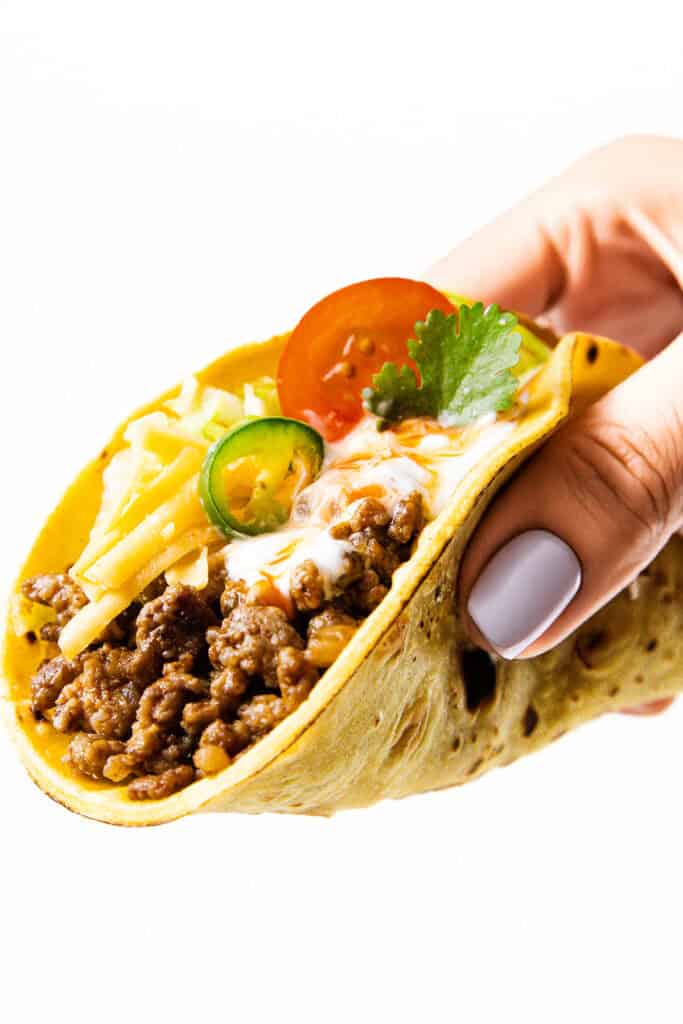 A hand holding a ground beef taco filled with homemade seasoning, shredded cheese, lettuce, tomato, jalapeño slices, cilantro, and sour cream, against a white background.