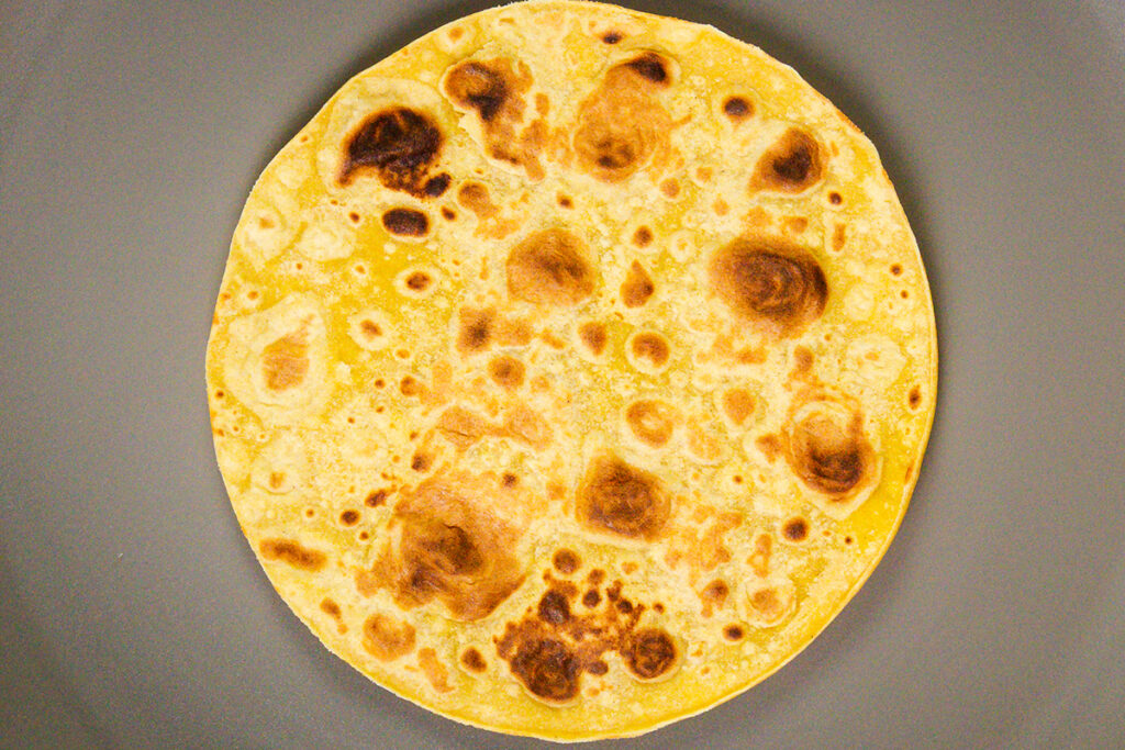 A close-up of a single, round, golden-brown flatbread with unevenly toasted spots, placed on a plain gray surface—perfect for serving smoky ground beef tacos.