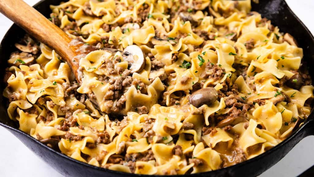 A skillet filled with beef stroganoff, featuring wide egg noodles, ground beef, sliced mushrooms, and a creamy sauce, garnished with chopped herbs and stirred with a wooden spoon.