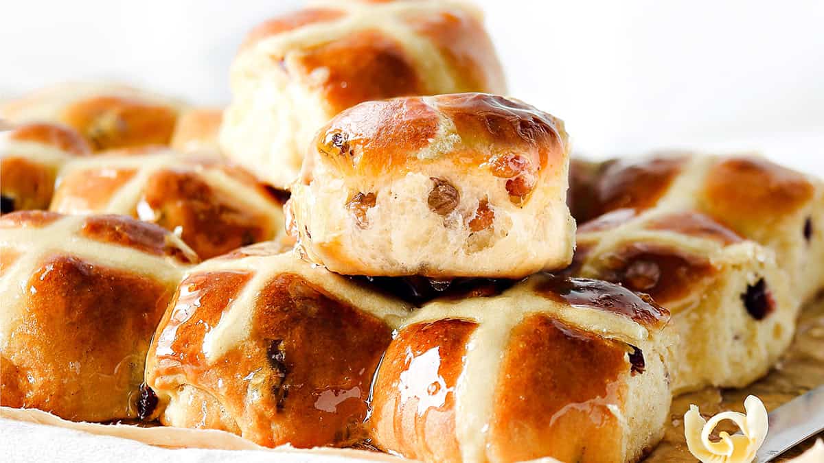 A close-up of a stack of glossy, freshly baked hot cross buns with raisins and white icing crosses invites your taste buds to brunch. Arranged on parchment paper, these delightful treats with a small flower on the side are perfect for any brunch recipe collection.