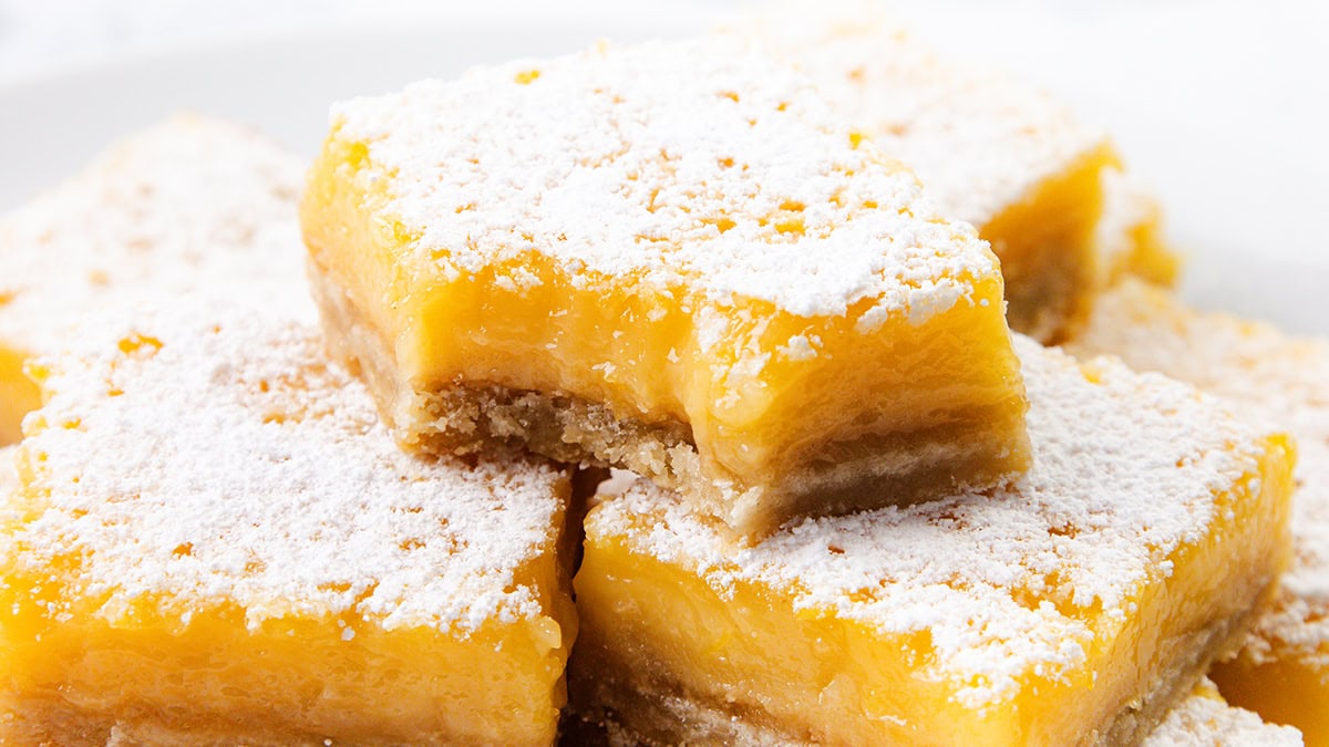 Close-up of lemon bars stacked on a plate, topped with powdered sugar. One bar has a bite taken out, revealing the creamy yellow lemon filling and buttery crust—a classic nod to beloved 1980s desserts.