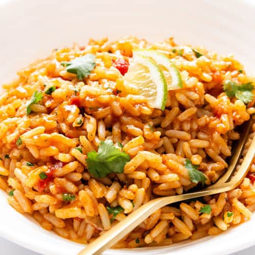 A white bowl filled with flavorful Mexican Rice, garnished with fresh cilantro and lemon wedges, with a gold fork resting on the side of the bowl.