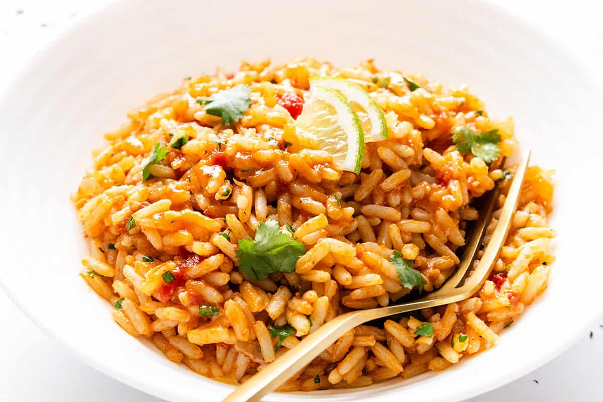 A white bowl filled with flavorful Mexican Rice, garnished with fresh cilantro and lemon wedges, with a gold fork resting on the side of the bowl.