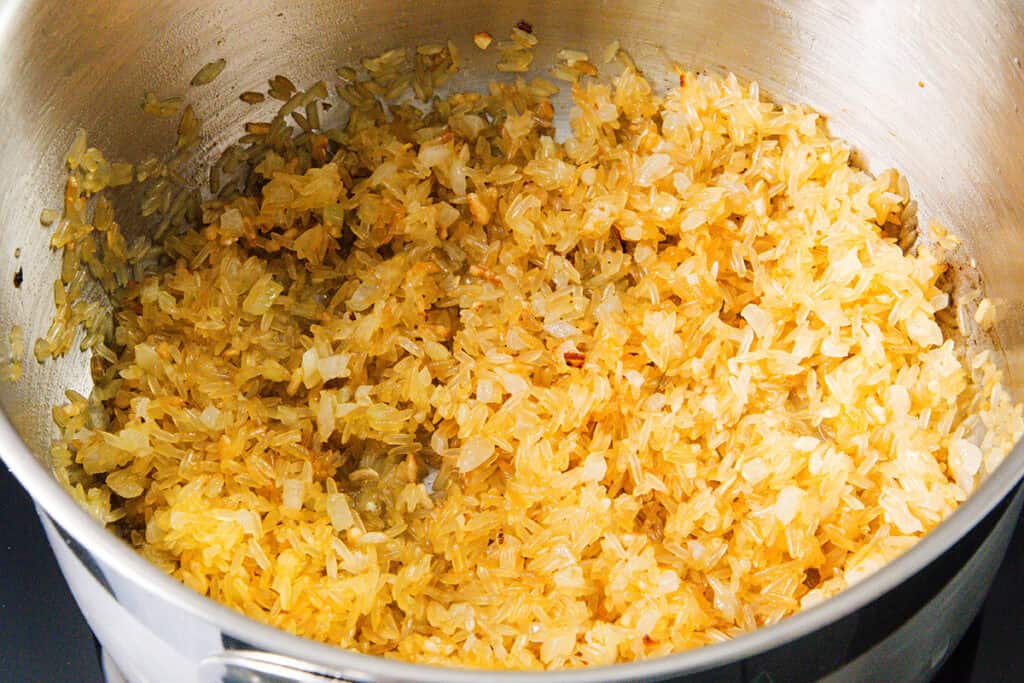 Chopped onions and rice are being sautéed in a stainless steel pot for Mexican Rice, turning golden brown as they cook.