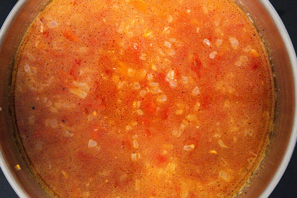 A close-up view of a pot filled with a simmering orange-red tomato-based sauce, perfect for an authentic Mexican Rice recipe, with visible chunks of onions and tomatoes.