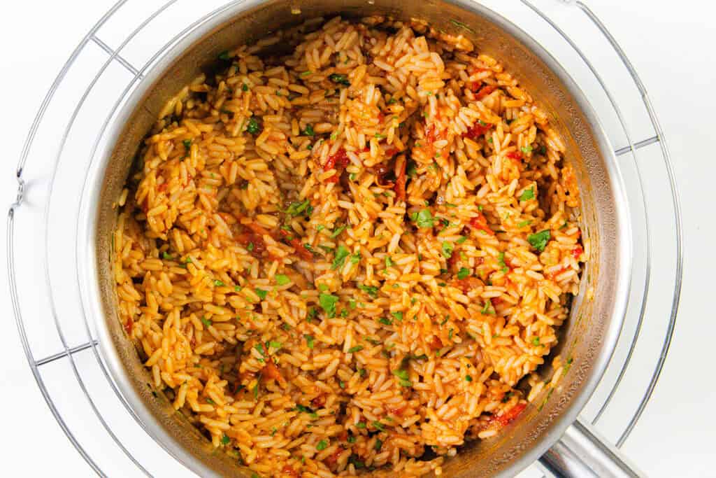 A stainless steel pot filled with fluffy Mexican Rice, orange-red in color and seasoned with bits of tomato and fresh herbs, sits garnished atop a round wire cooling rack.