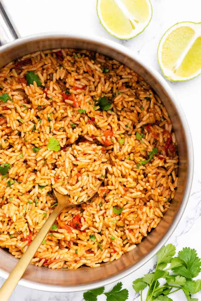 A pot of authentic Mexican Rice garnished with cilantro, with a golden spoon inside. Fresh lime halves and cilantro leaves are placed nearby on a white surface.