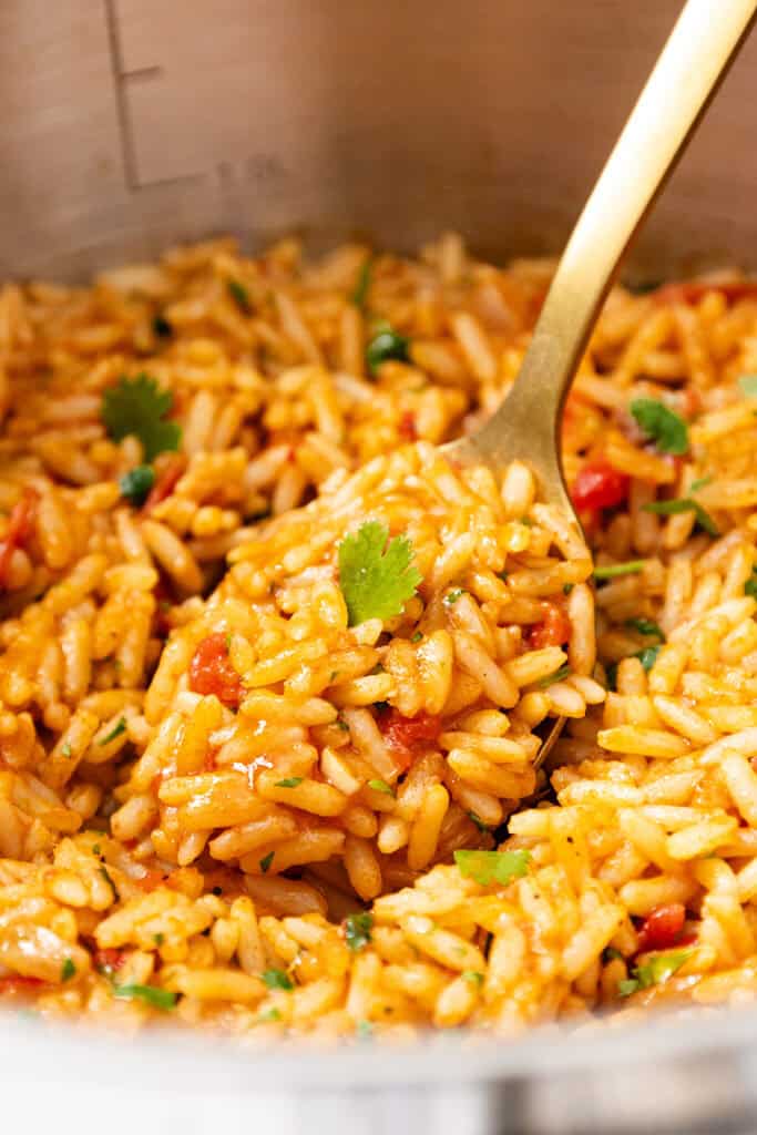 A close-up of cooked orzo pasta mixed with tomato sauce and herbs, stirred with a gold-colored fork in a stainless steel pot, reminiscent of the vibrant flavors found in classic Mexican Rice dishes.