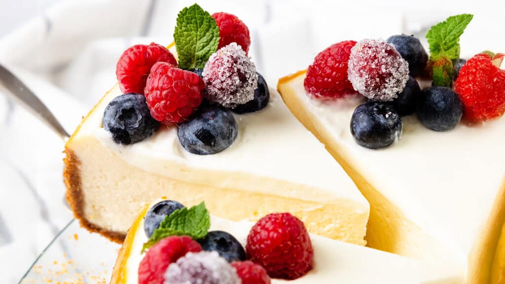 Slices of creamy cheesecake topped with fresh raspberries, blueberries, and mint leaves. The berries are slightly dusted with sugar, adding a decorative touch. The background includes a soft, white cloth.