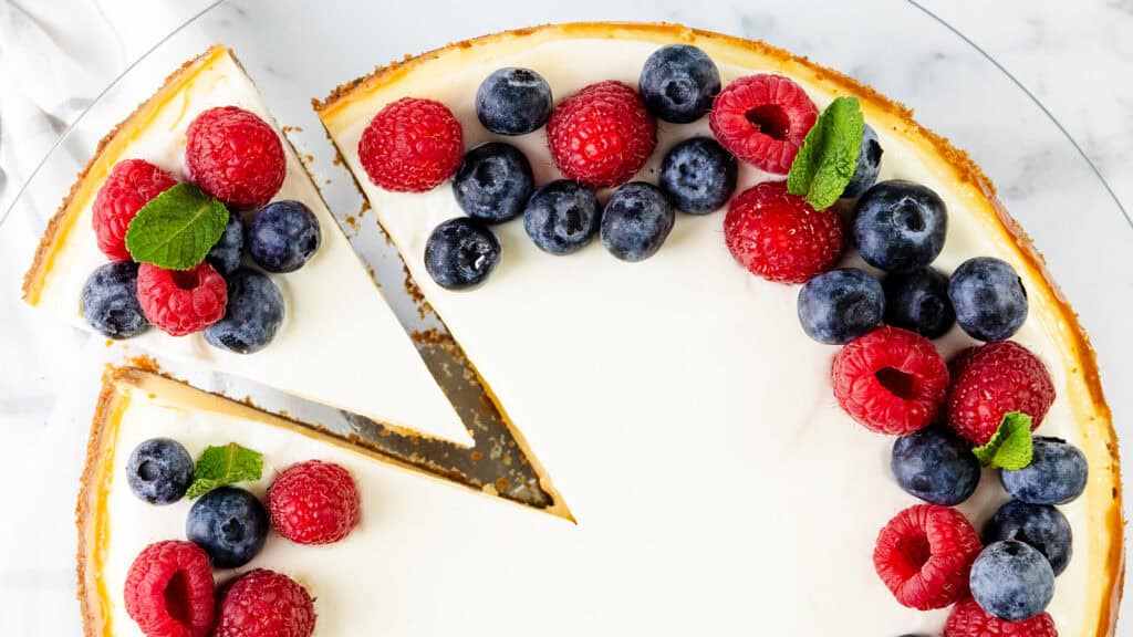 A cheesecake topped with fresh raspberries, blueberries, and mint leaves—perfect for Memorial Day Recipes. One slice is slightly separated from the rest of the cake, revealing its creamy texture as it sits elegantly on a glass plate.