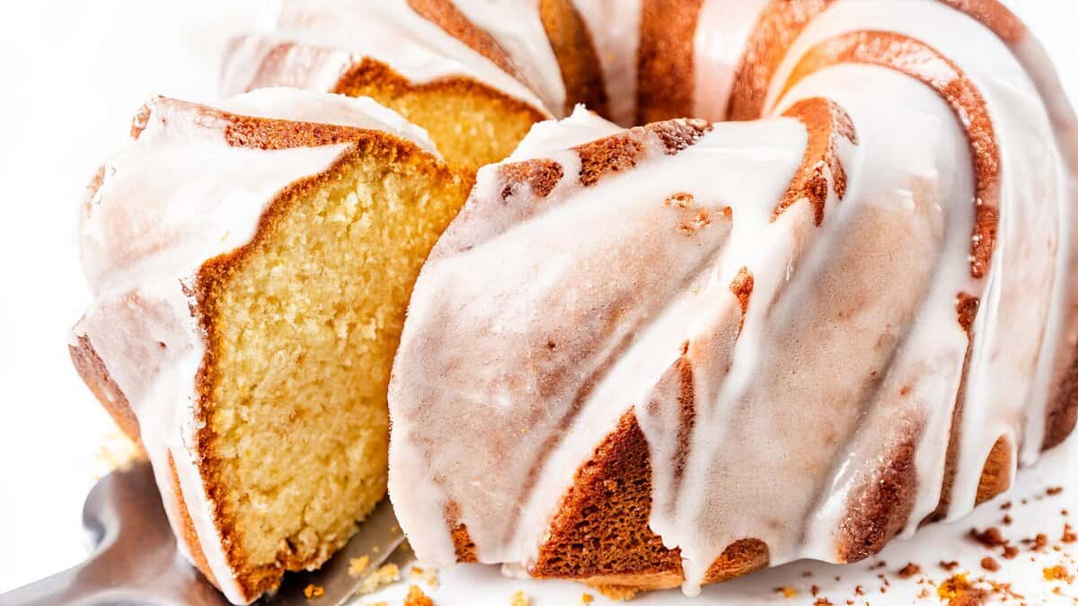 Close-up of a glazed bundt cake with a slice cut out, revealing a moist yellow interior. The cake is covered with a shiny, white icing, and is served on a platter. Crumbs are visible near the base, adding texture to the scene.