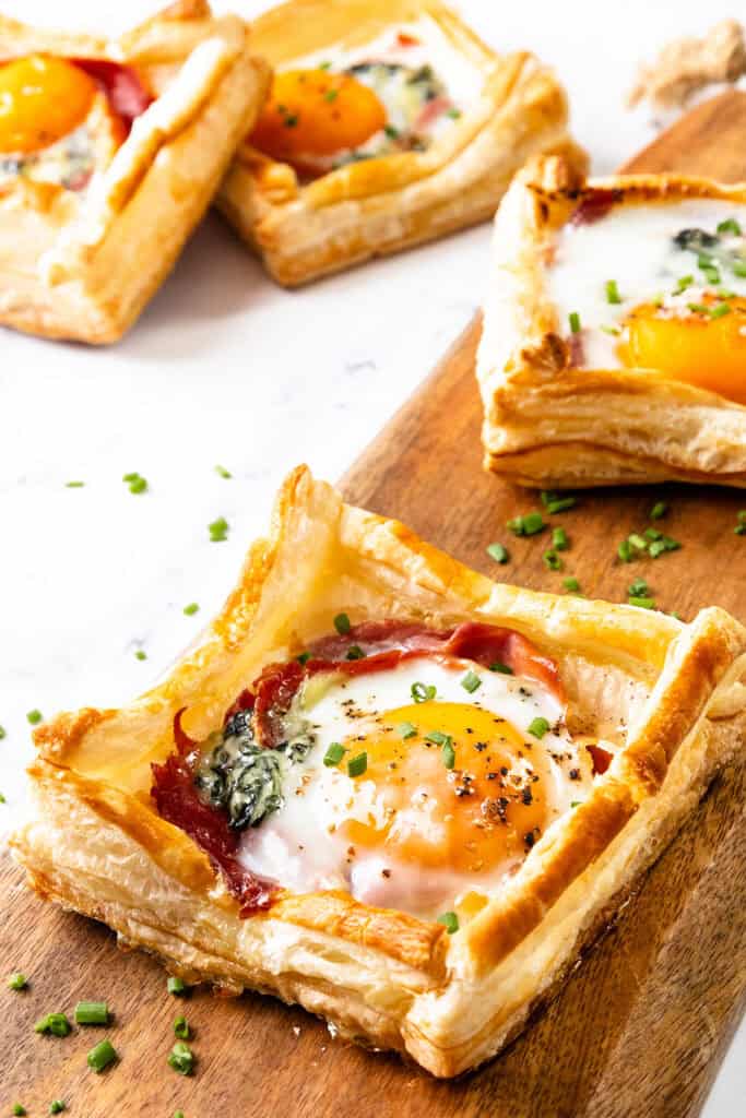 Square puff pastry breakfast tarts topped with sunny-side-up eggs, spinach, prosciutto, and chopped chives are arranged on a wooden board. The tarts have golden, flaky edges and a colorful, appetizing appearance.