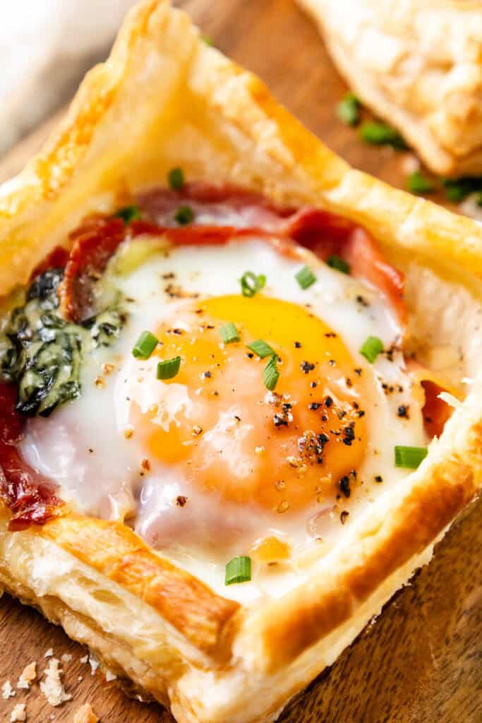 This Puff Pastry Breakfast features a golden, flaky square topped with a baked egg, cooked spinach, savory prosciutto, and chopped chives, served on a wooden surface.