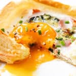 A flaky puff pastry breakfast square filled with prosciutto, spinach, cheese, and a sunny-side-up egg with a runny yolk, garnished with chopped chives on a white plate.