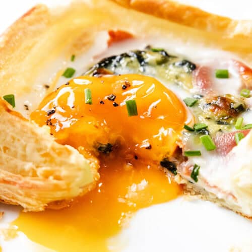A flaky puff pastry breakfast square filled with prosciutto, spinach, cheese, and a sunny-side-up egg with a runny yolk, garnished with chopped chives on a white plate.