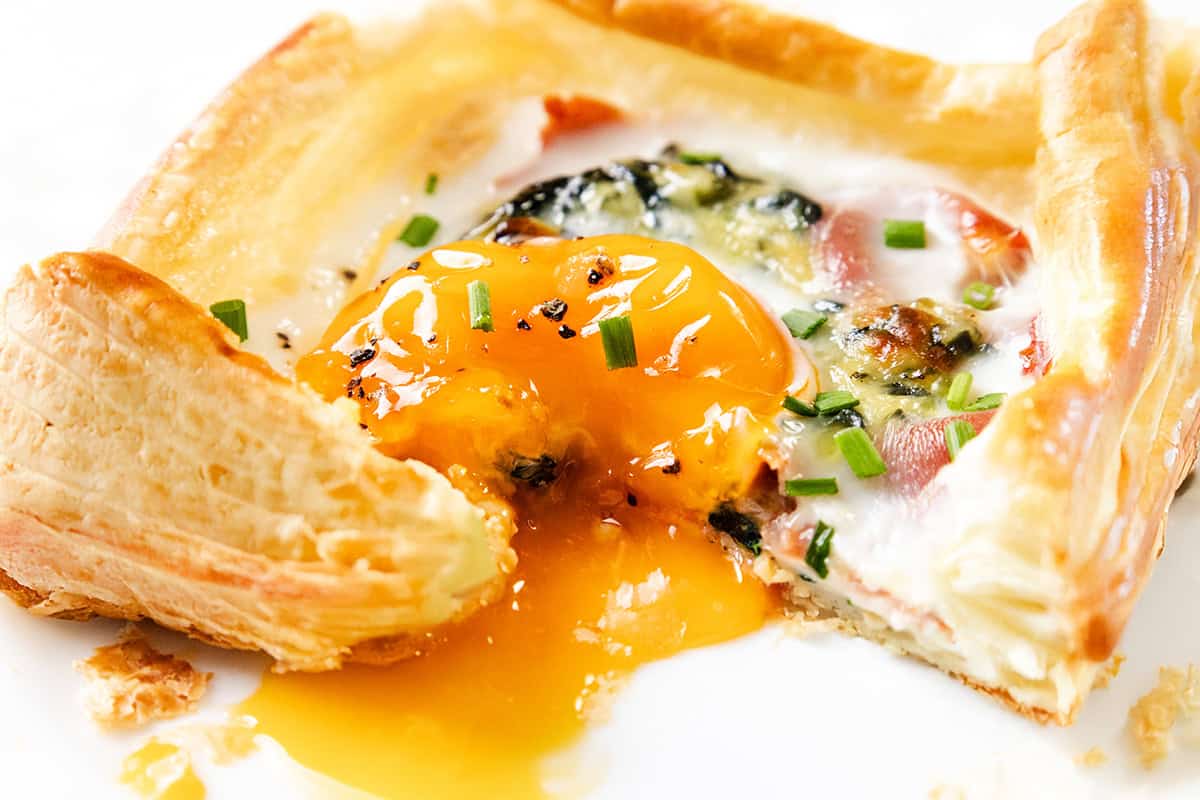 A flaky puff pastry breakfast square filled with prosciutto, spinach, cheese, and a sunny-side-up egg with a runny yolk, garnished with chopped chives on a white plate.