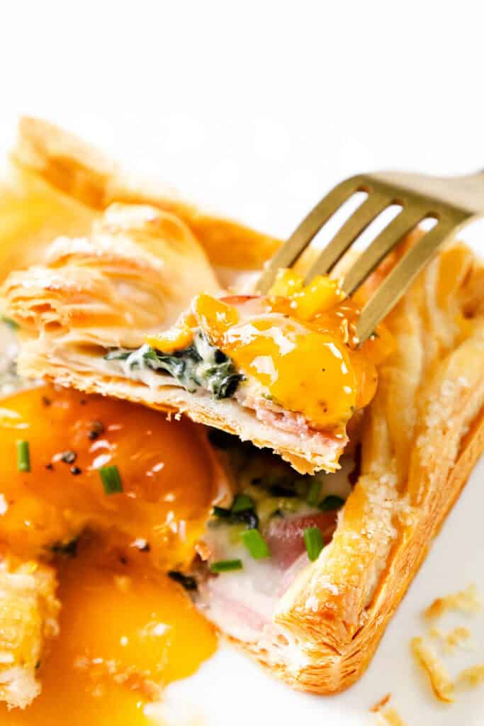 A close-up of a flaky puff pastry breakfast filled with prosciutto, spinach, and a runny egg yolk, with a fork lifting a bite-sized piece. The pastry is garnished with chopped chives.
