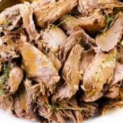 A close-up of a plate filled with tender, juicy slices of slow-cooked leg of lamb garnished with fresh thyme sprigs. The meat is golden brown and appears to be flavorful and moist.