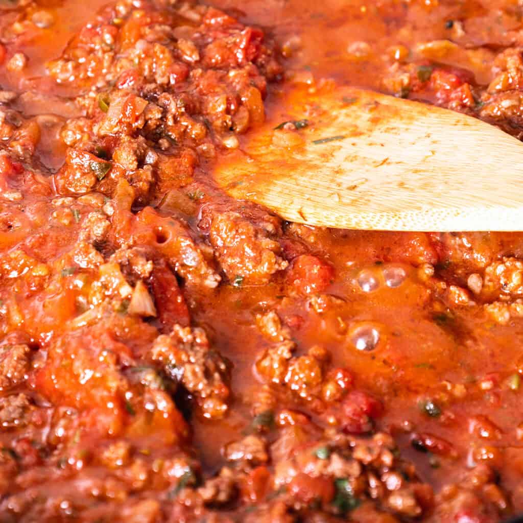 Close-up of a wooden spoon stirring a rich, chunky tomato and meat sauce. The sauce has visible pieces of ground meat, diced tomatoes, and herbs, all simmering together, creating a hearty texture in a vibrant red-orange hue.