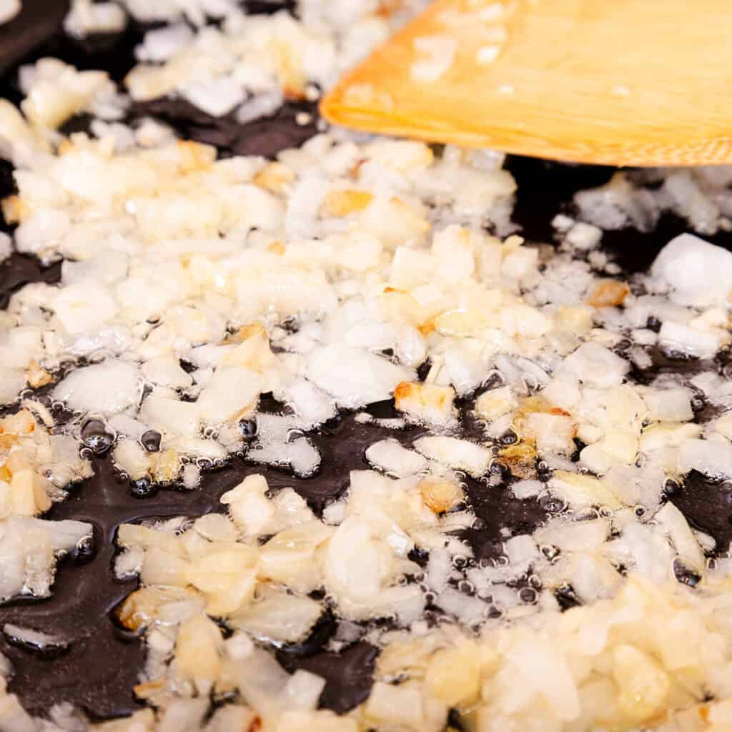 Chopped onions being sautéed in oil on a black skillet, stirred with a wooden spatula. The onions are partially translucent and golden brown, indicating they are starting to caramelize.