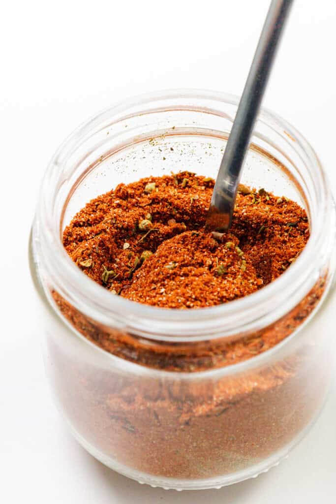 A glass jar filled with a reddish-brown homemade seasoning, perfect for smoky tacos, with a metal spoon partially submerged in the blend. The jar is set against a plain white background.