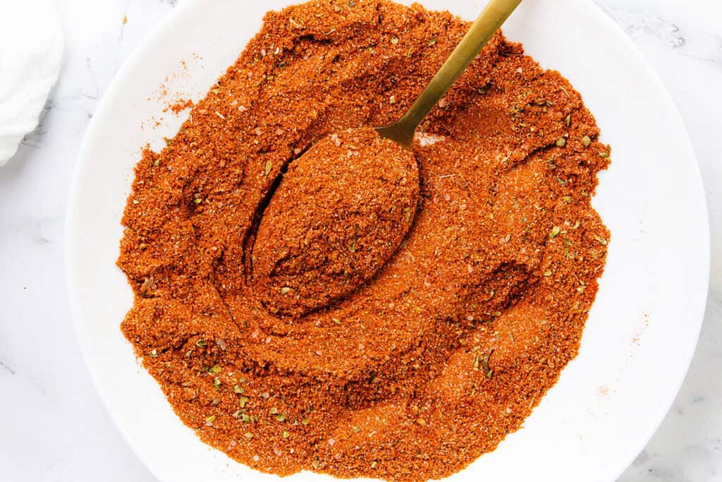 A white plate filled with a reddish-brown homemade seasoning blend, with a gold spoon scooping some of the mixture. Perfect for smoky tacos or ground beef tacos, the blend contains visible herbs and spices on a white marble surface.