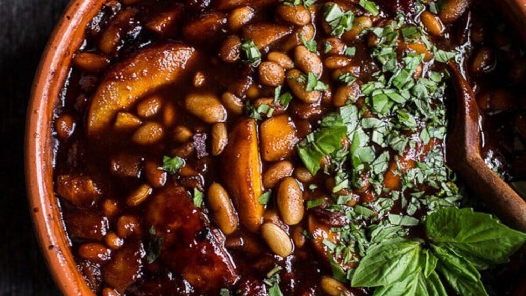 A close-up of a bowl of baked beans with sliced sausages and fresh herbs on top, garnished with chopped parsley or cilantro—a classic comfort food inspired by Southern cooking, with a wooden spoon resting inside.