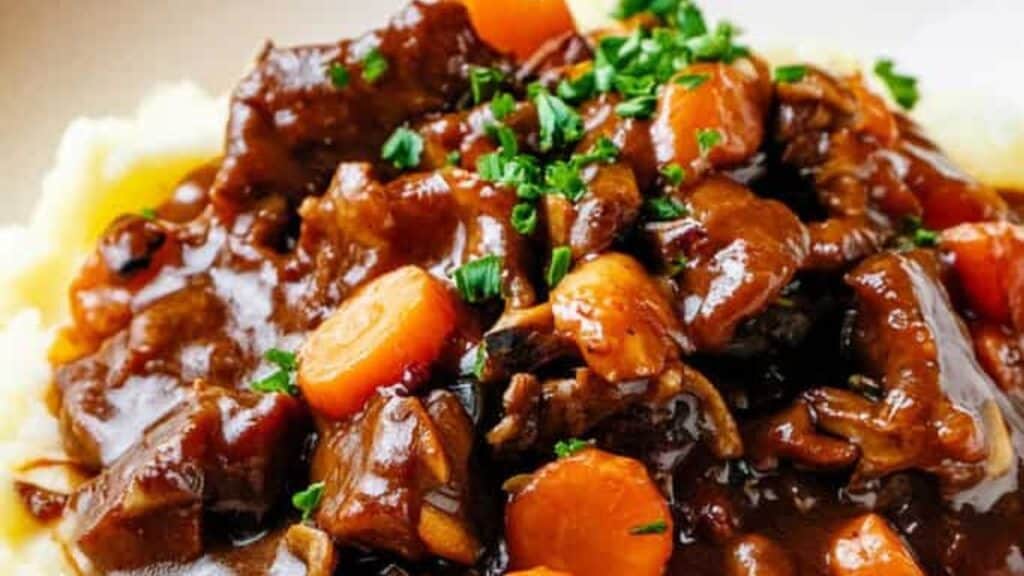 A close-up of beef stew with chunks of beef, sliced carrots, and rich brown gravy, garnished with chopped parsley, served over mashed potatoes.