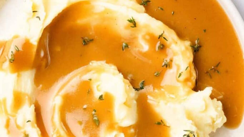 A close-up of creamy mashed potatoes topped with brown gravy and garnished with small green herb leaves on a white plate.