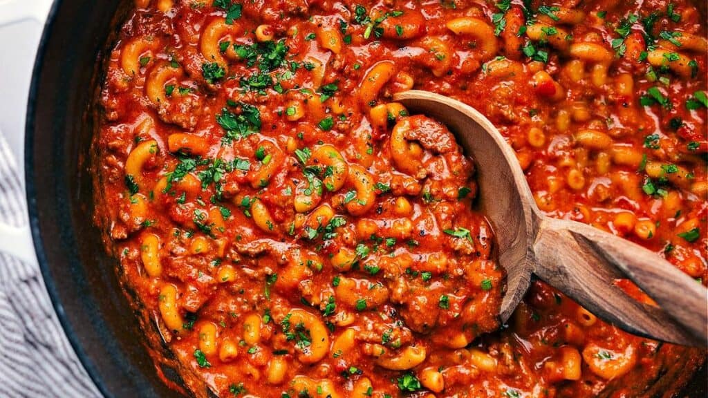 A close-up of American goulash, showing elbow macaroni, ground beef, and tomato sauce garnished with chopped parsley, being stirred with a wooden spoon in a pot.