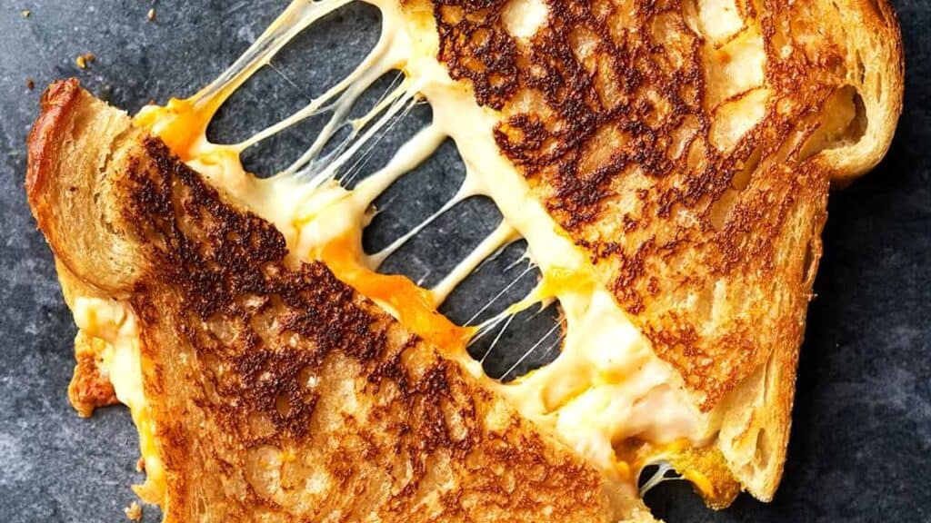 A grilled cheese sandwich cut in half, with melted cheese stretching between the halves, on a dark surface. The bread is golden brown and toasted.