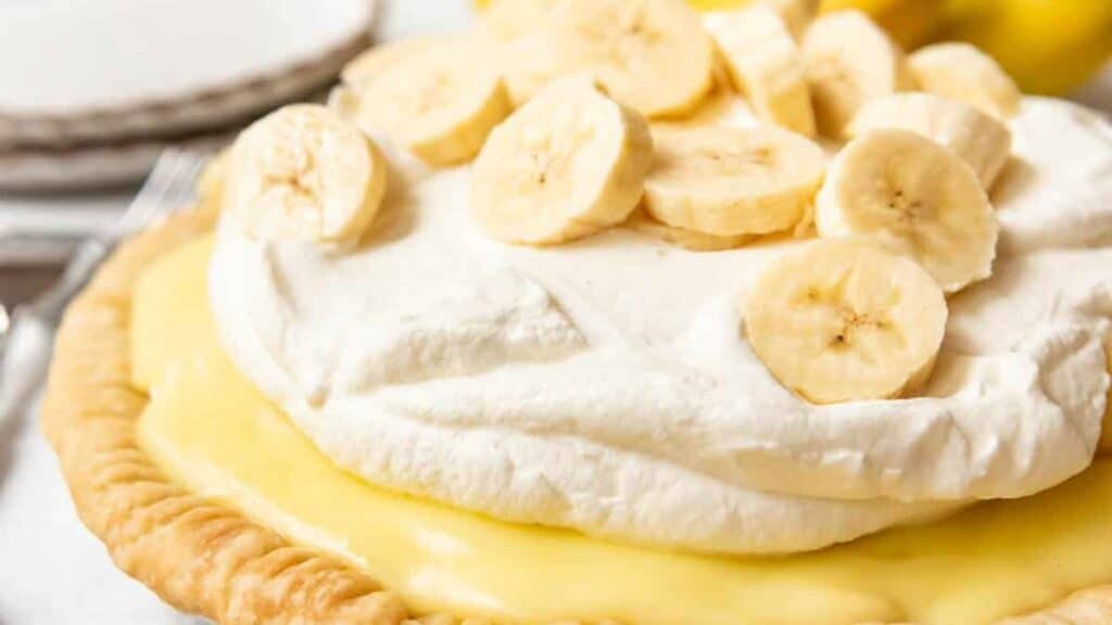 A close-up of a banana cream pie topped with whipped cream and banana slices, showing the creamy yellow filling and flaky pie crust.