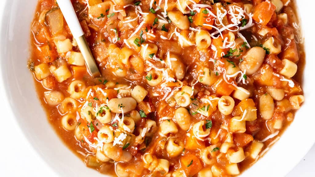 A close-up of a bowl of pasta e fagioli soup, a classic Italian dish with ditalini pasta, white beans, tomatoes, carrots, shredded cheese, and herbs—perfect for those who love authentic Italian recipes. A spoon rests in the bowl.