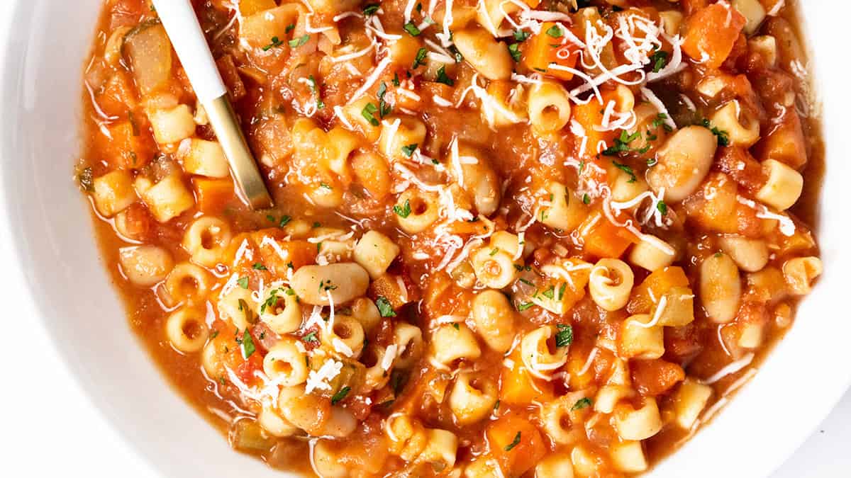 A close-up of a bowl of pasta e fagioli soup, a classic Italian dish with ditalini pasta, white beans, tomatoes, carrots, shredded cheese, and herbs—perfect for those who love authentic Italian recipes. A spoon rests in the bowl.