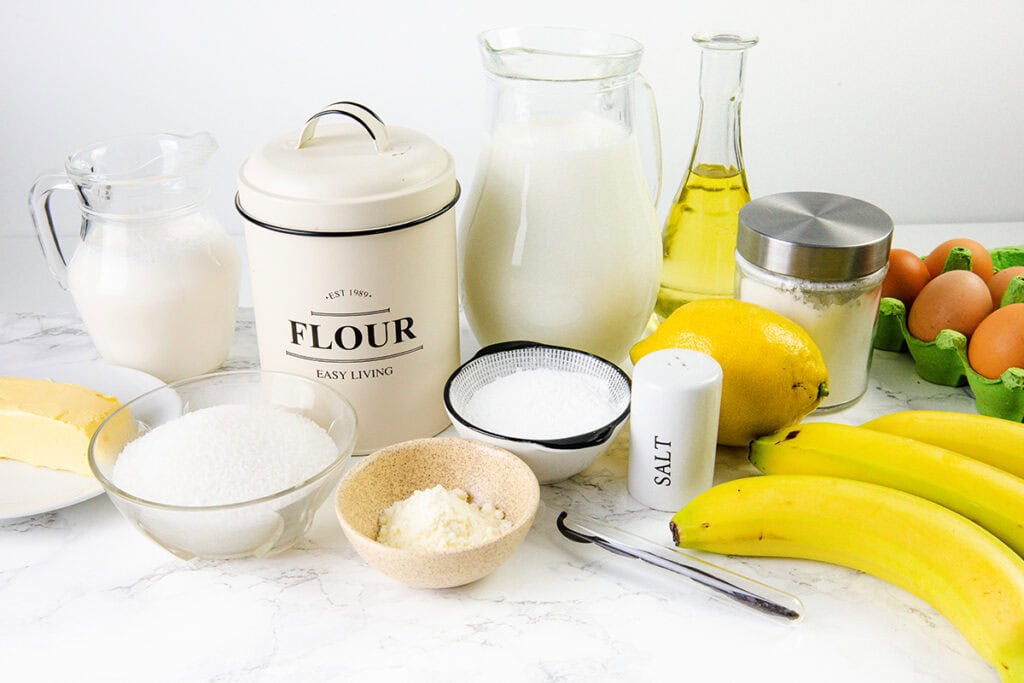 A variety of baking ingredients for an old-school pie on a white surface, including flour, milk, oil, sugar, eggs in a carton, bananas, vanilla pod, lemon, salt shaker, butter, and cream in various containers.