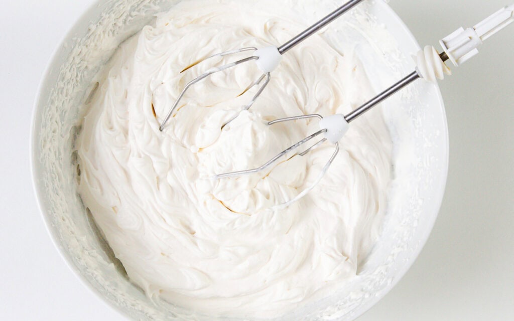 A close-up of a white mixing bowl filled with whipped cream, with two metal electric mixer beaters resting on top—perfectly prepped for an old-school Banana Cream Pie.