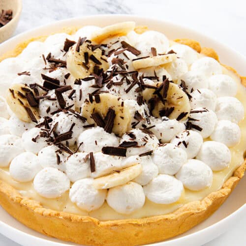 An Old-School banana cream pie topped with whipped cream, banana slices, and chocolate shavings on a white plate sits on a marble surface. A bowl of chocolate shavings is in the background.