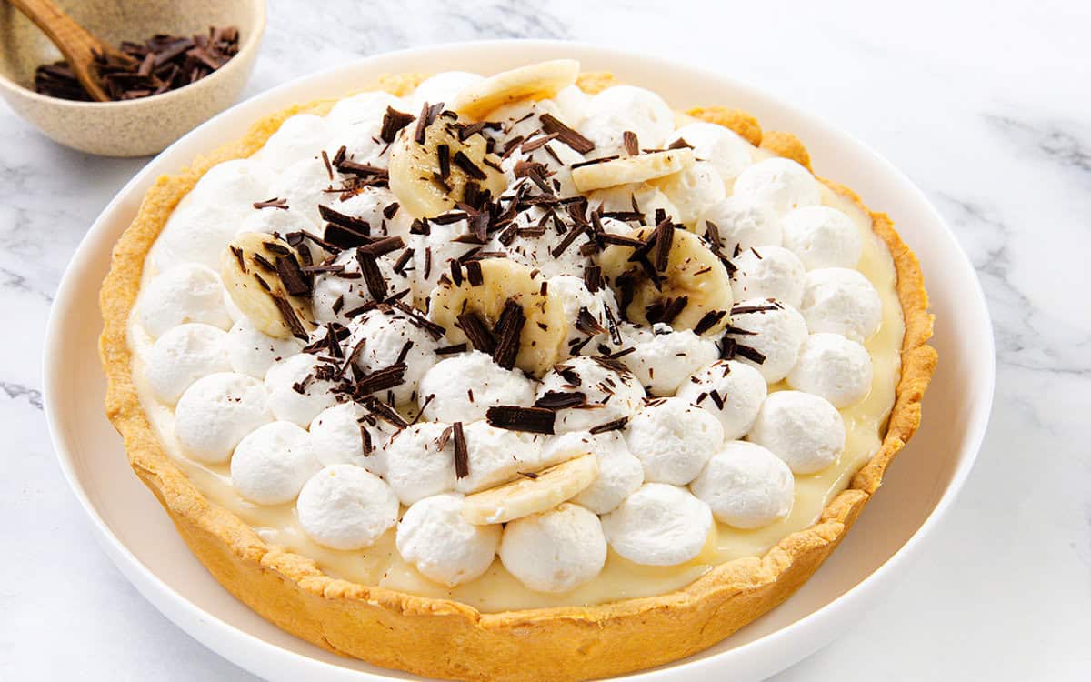An Old-School banana cream pie topped with whipped cream, banana slices, and chocolate shavings on a white plate sits on a marble surface. A bowl of chocolate shavings is in the background.