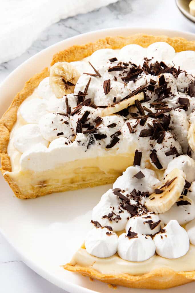 A classic Banana Cream Pie with a slice cut out, topped with whipped cream, banana slices, and chocolate shavings on a white plate—a true old-school dessert favorite.