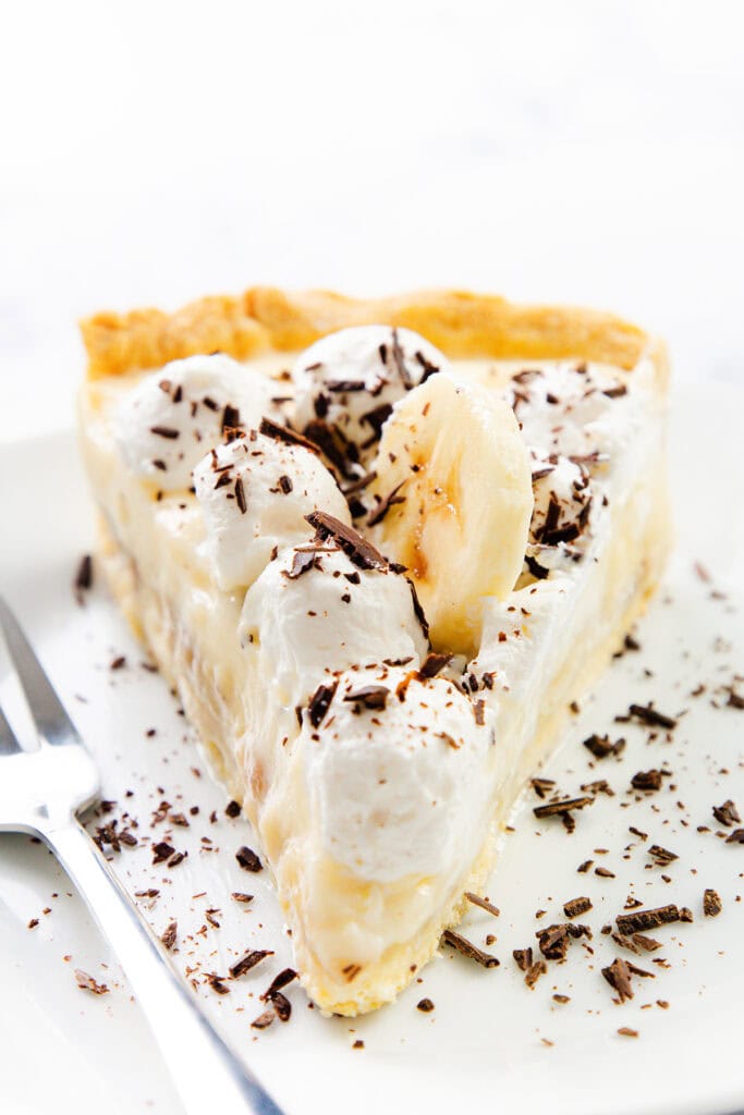 A slice of old-school banana cream pie topped with whipped cream, banana slices, and chocolate shavings on a white plate, with a fork beside it.
