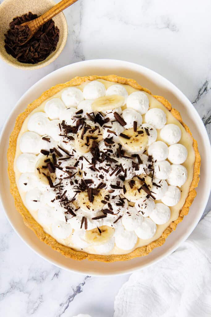 An old-school banana cream pie topped with whipped cream, banana slices, and chocolate shavings sits on a white plate, with a bowl of chocolate shavings and a spoon nearby on a marble surface.