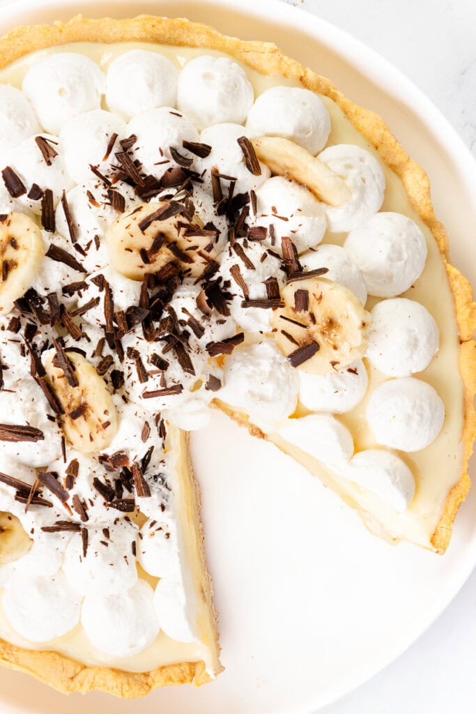 A classic Banana Cream Pie topped with whipped cream, banana slices, and chocolate shavings sits on a white plate, one slice missing—a dreamy banana dessert and true old-school pie favorite.